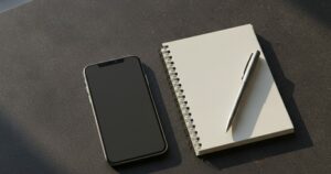 Martwa natura: smartfon lezacy ekranem do gory na ciemnym blacie, obok notes i dlugopis, chlodne swiatlo. Ekran pusty, bez interfejsu. BEZ ludzi, BEZ twarzy, BEZ napisow i liter.