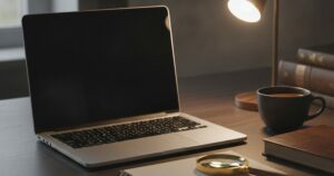Martwa natura: otwarty laptop na ciemnym biurku, ekran wygaszony, obok notes, lupa i kubek kawy, wieczorne swiatlo lampki. BEZ ludzi, BEZ twarzy, BEZ napisow i liter, BEZ interfejsu na ekranie.
